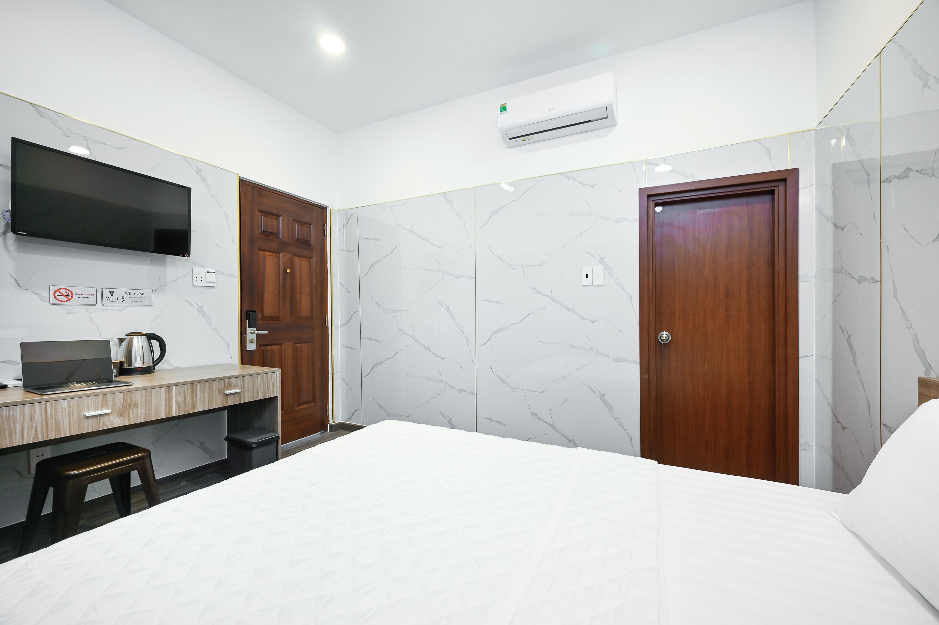 A hotel room with a bed, a desk with a laptop and a coffee maker, a TV mounted on the wall, and marble-patterned walls.