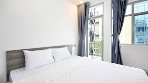 A neatly made bed with white bedding and a wooden headboard in a hotel room with large windows and grey curtains. - Yoko Airport Saigon Hotel (Ho Chi Minh City)