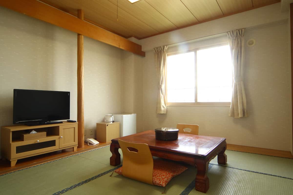 Une pièce de style japonais traditionnel, avec un sol recouvert de tatamis, une table en bois, deux chaises, un téléviseur posé sur un meuble en bois et une fenêtre avec des rideaux.