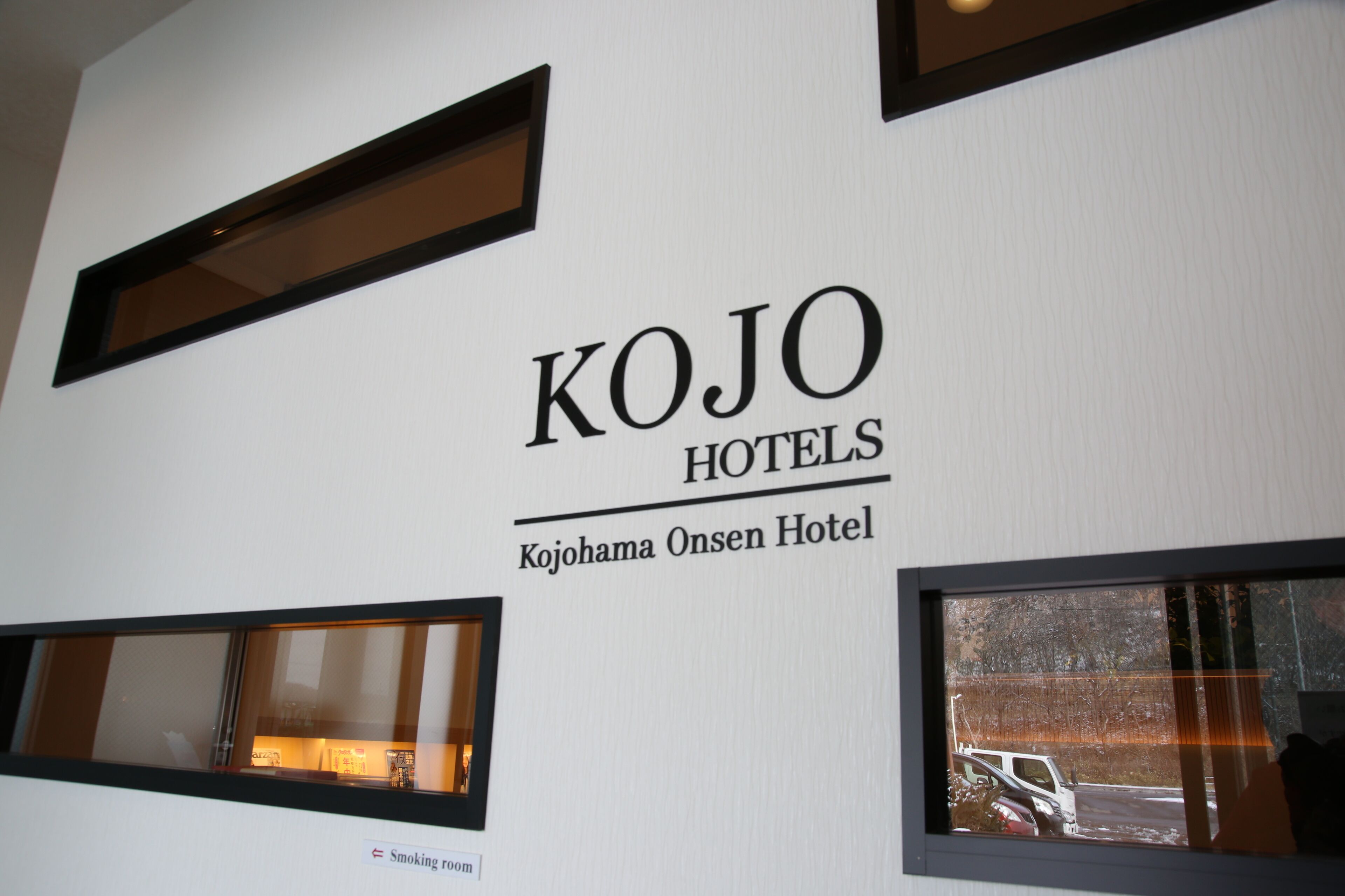 Señal de KOJO HOTELS, concretamente del Kojohama Onsen Hotel, con vista a un aparcamiento a través de una ventana.