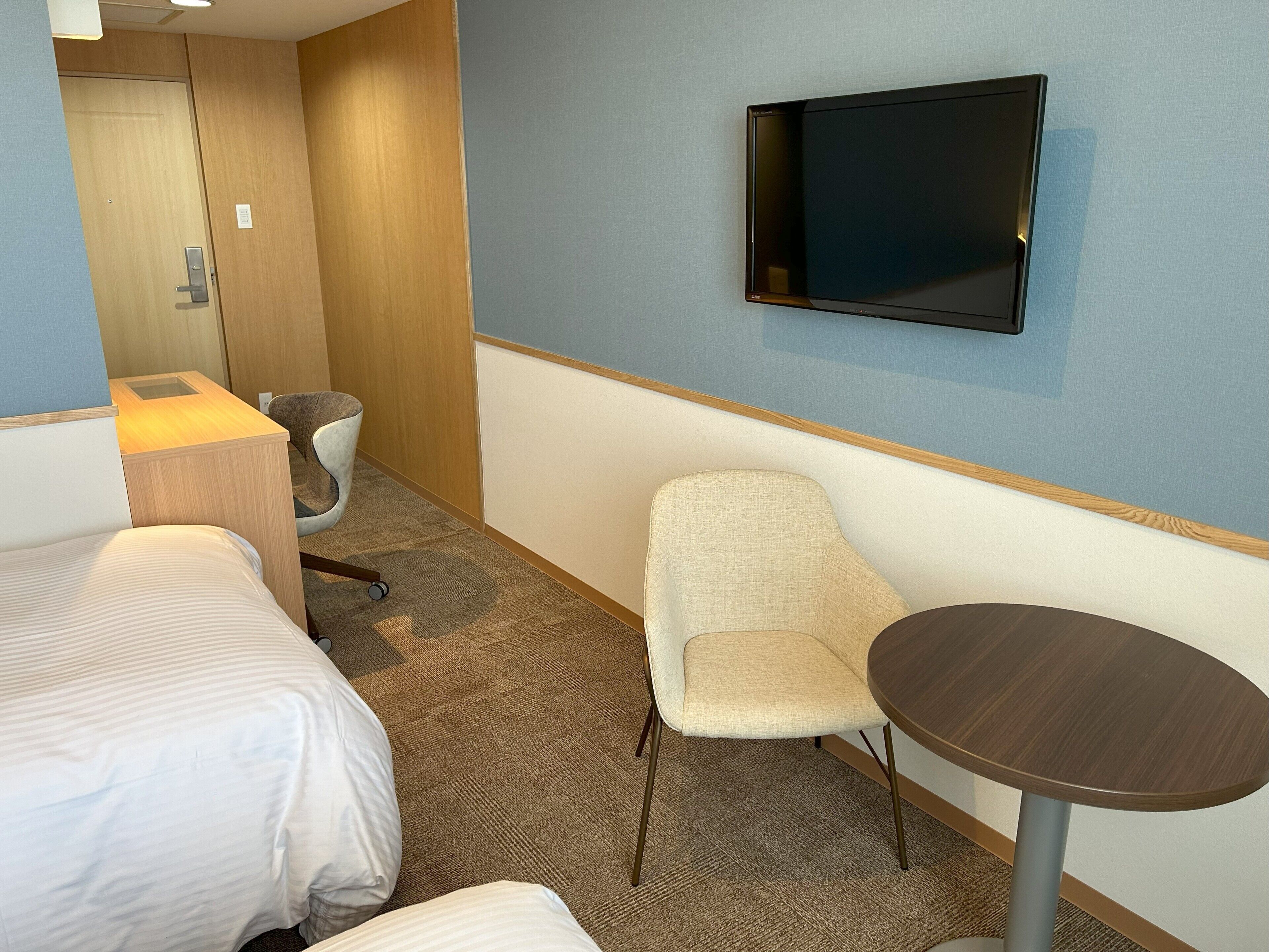 Een hotelkamer met een bed, een stoel, een klein tafeltje, een aan de muur bevestigde televisie en een deur.