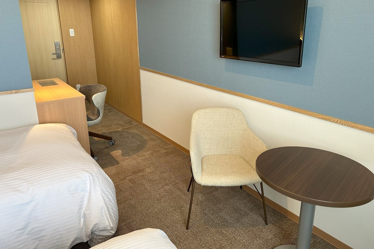 Een hotelkamer met een bed, een stoel, een klein tafeltje, een aan de muur bevestigde televisie en een deur.