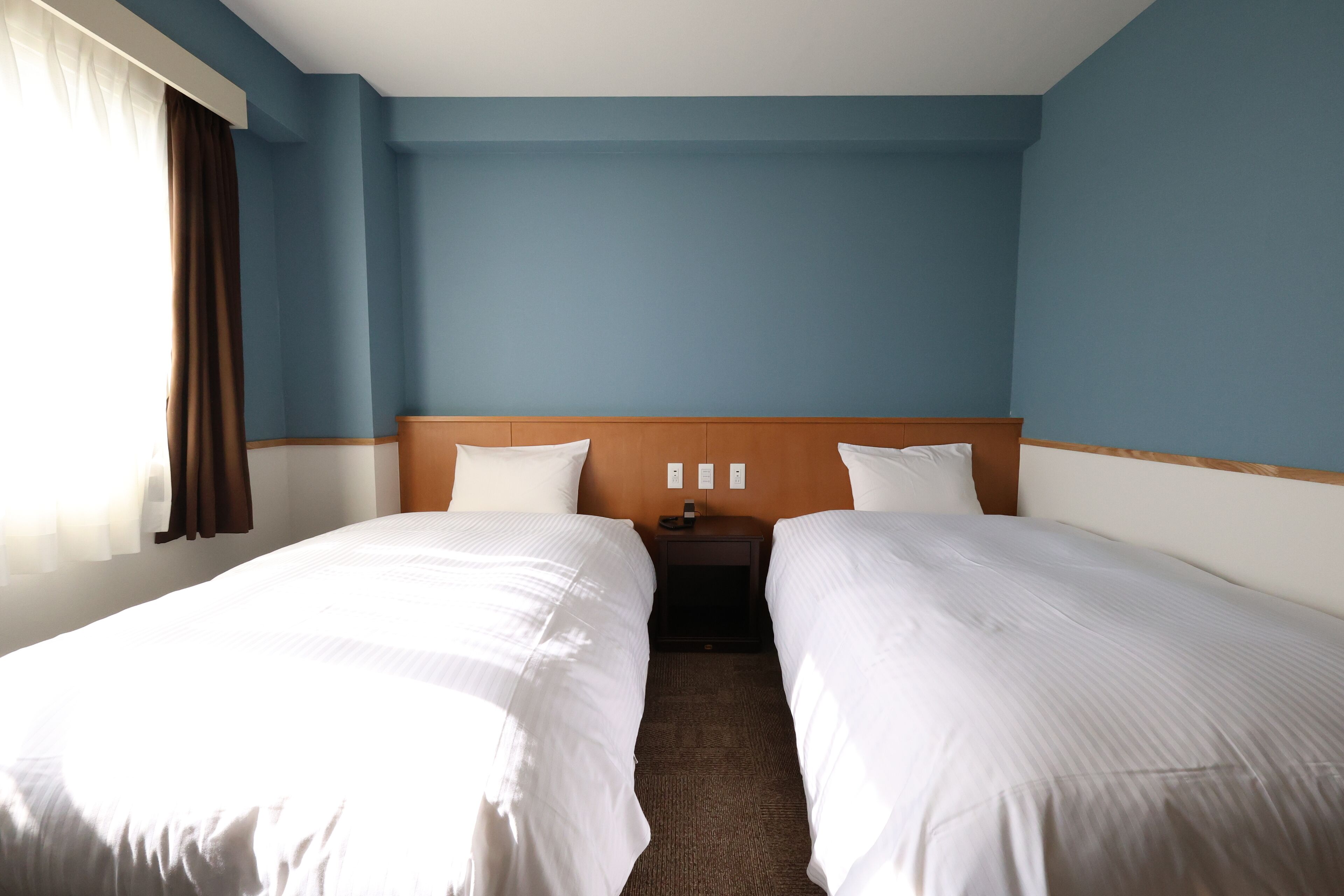 A hotel room with two beds, a wooden headboard, a nightstand, and a window with curtains.