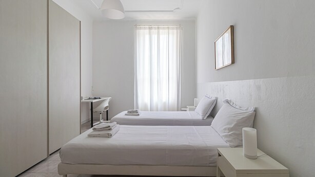 A minimalist hotel room with two beds, a desk, and a large window with sheer curtains. - Italianway   - Argonne (Milan)