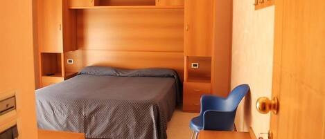 A bedroom with a bed, a desk, and a chair. There are wooden cabinets and a shelf above the bed.