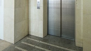 A modern elevator with a digital display and emergency buttons, located in a hallway with tiled walls and a striped floor. - At RLiS House Namba Minami (Osaka)