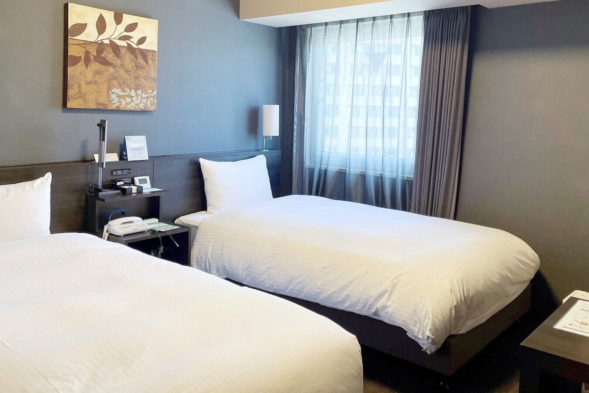 A hotel room with two beds, a desk with a lamp, a telephone, and a painting on the wall.