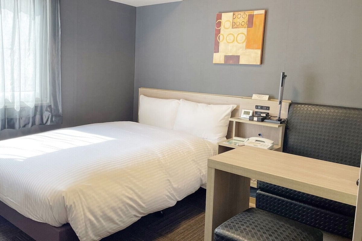 A hotel room with a bed, a desk, a chair, a nightstand, and a wall-mounted artwork.
