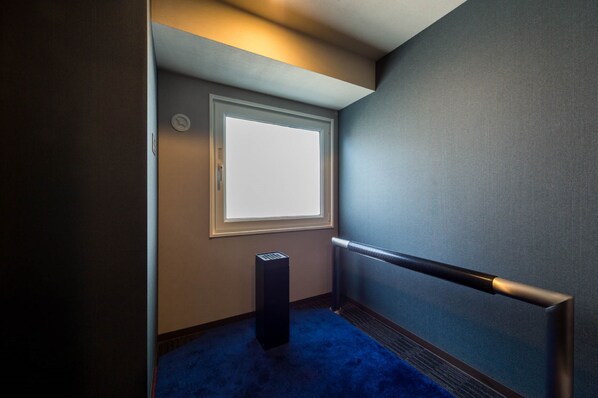 Um corredor estreito com carpete azul, uma porta com janela e um corrimão de metal.
