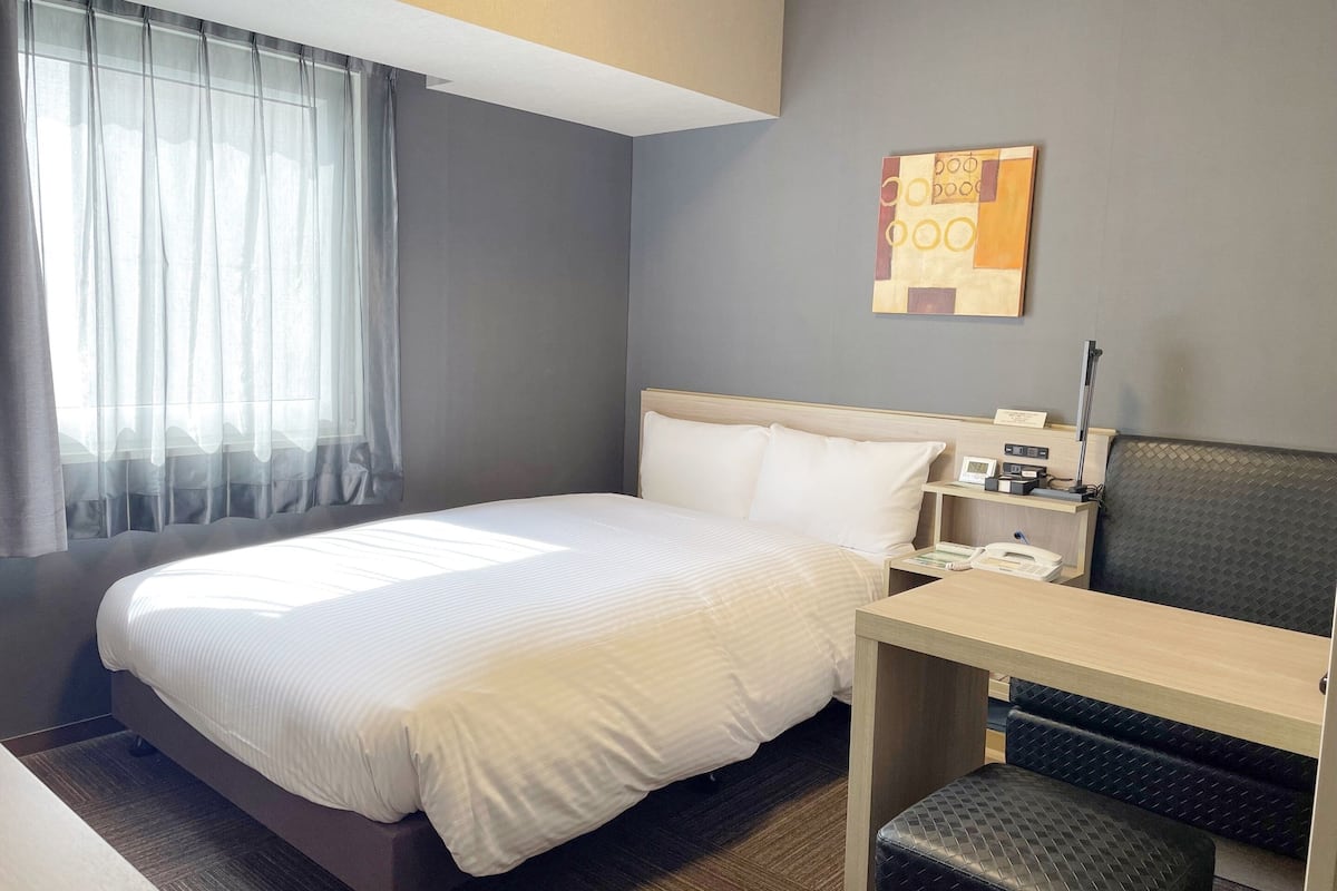 A hotel room with a bed, a desk, a chair, a nightstand, and a wall-mounted artwork.
