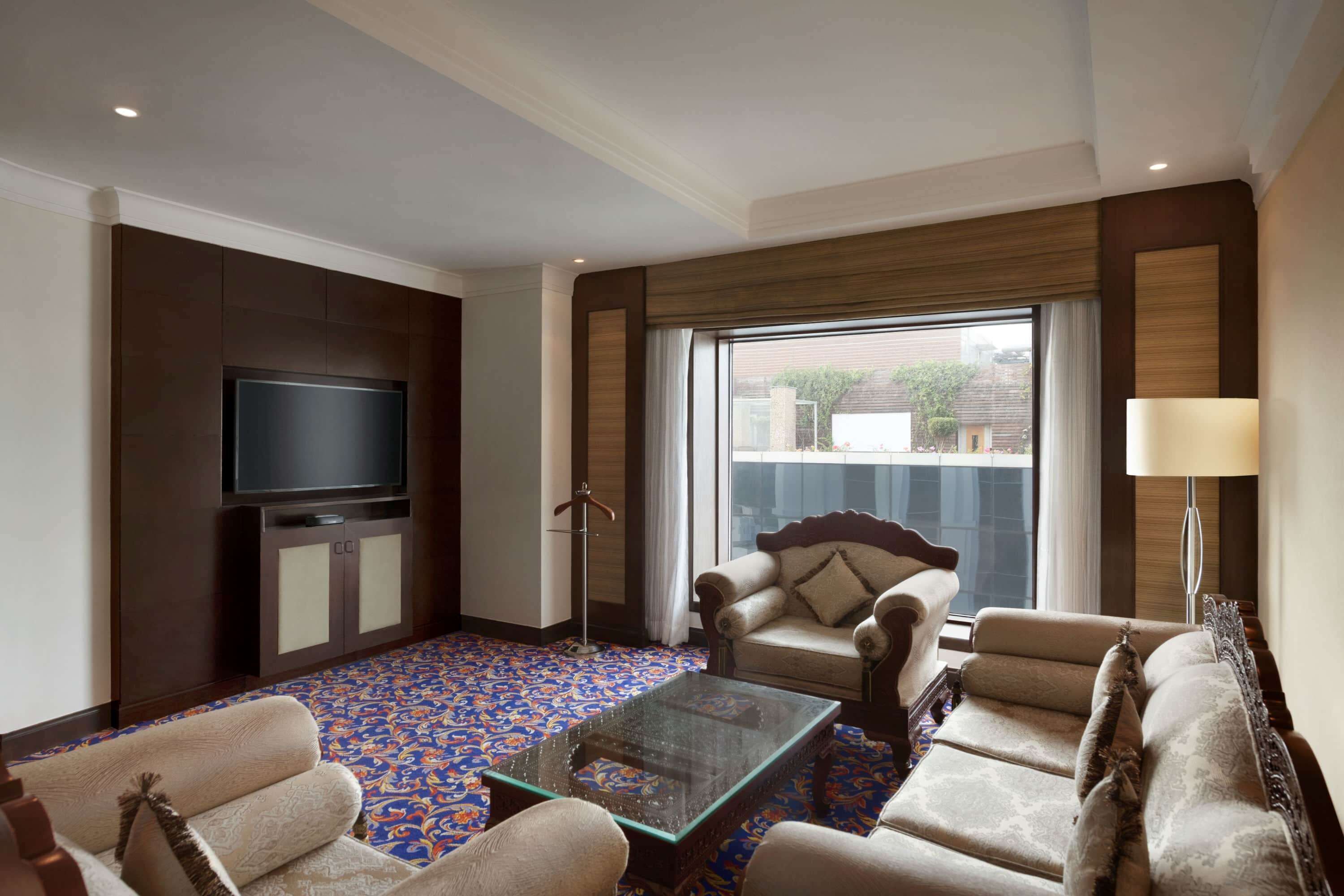 A hotel room with a large flat-screen TV, a sofa set, a glass coffee table, and a patterned carpet.