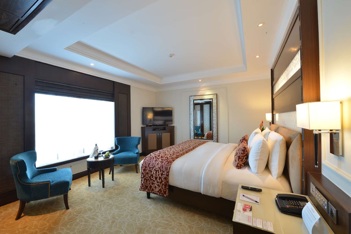 Une chambre d’hôtel avec un grand lit, un bureau avec un téléphone et des documents, une télévision, une chaise et une grande fenêtre.