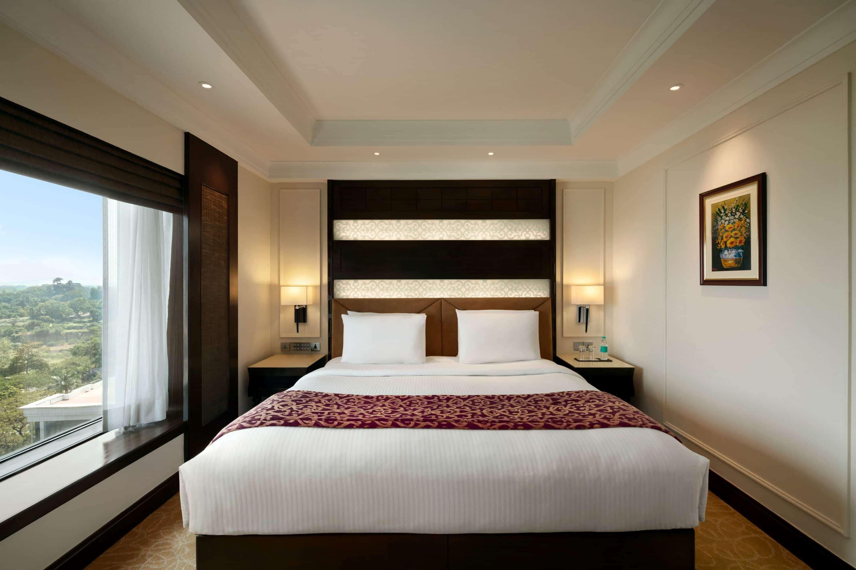 Una cama grande con cabecera, dos mesitas de noche, una ventana con cortinas e imagen enmarcada en la pared.