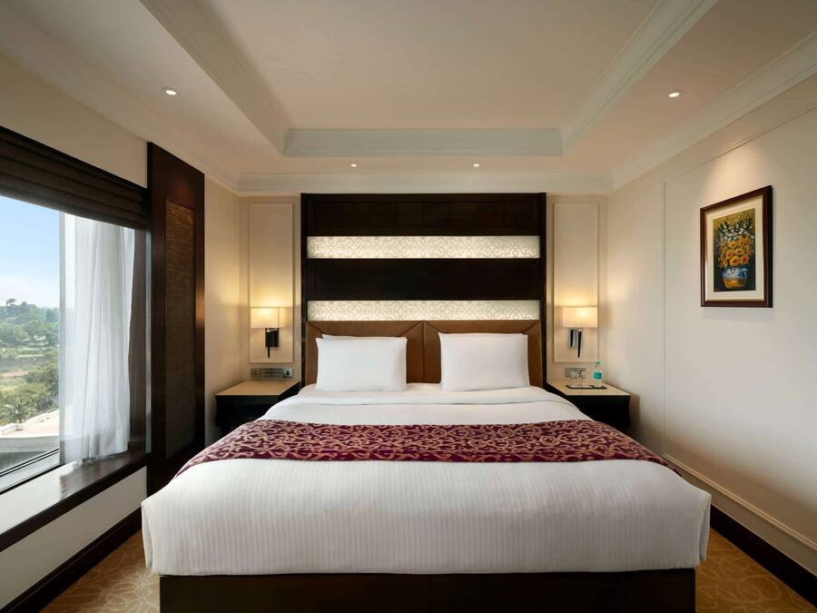 Una cama grande con cabecera, dos mesitas de noche, una ventana con cortinas e imagen enmarcada en la pared.
