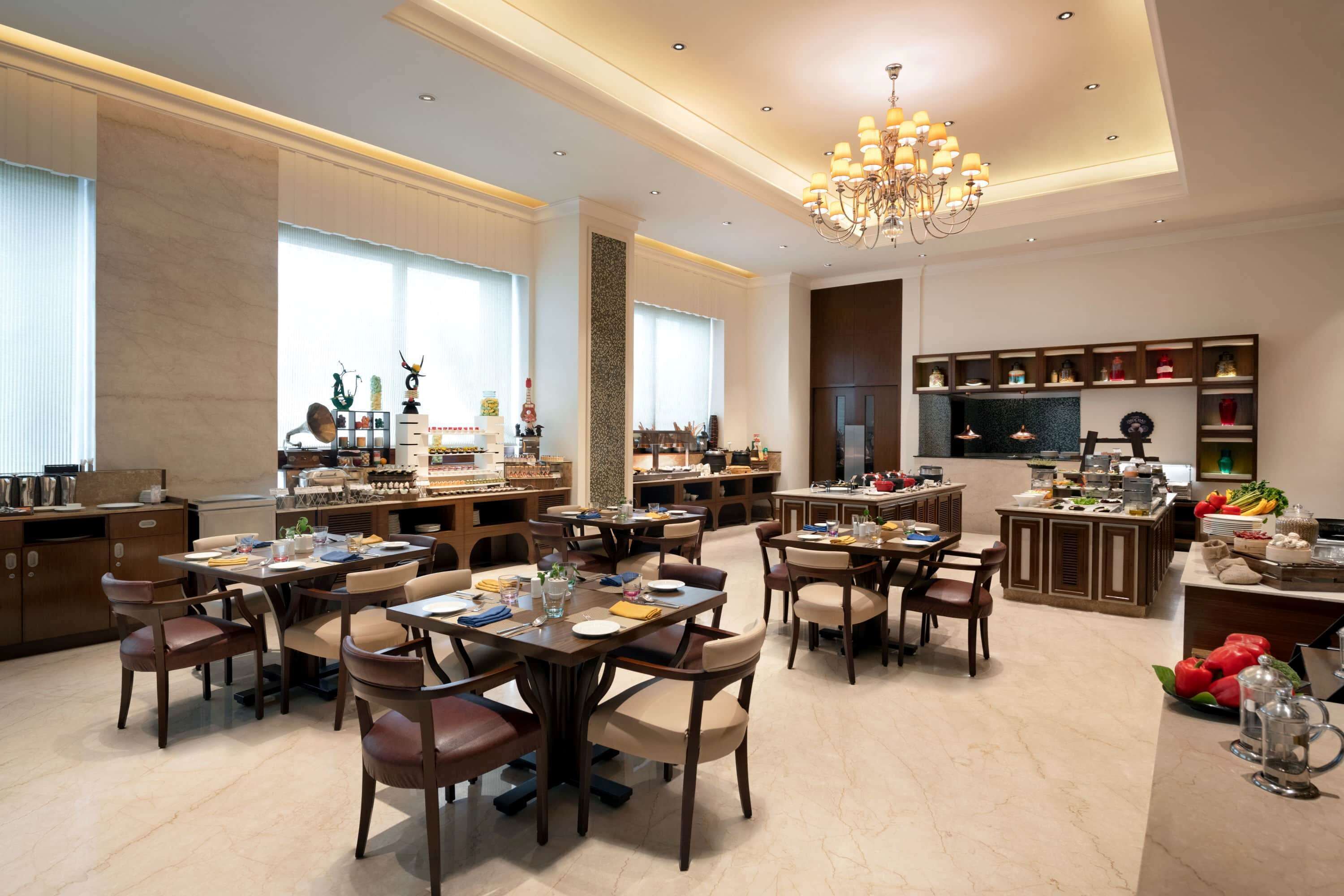 A spacious dining area with multiple tables and chairs, a central chandelier, and a kitchen area with various appliances and food displays.
