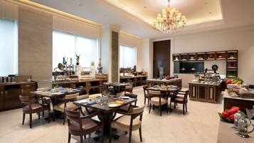 A spacious dining area with multiple tables and chairs, a central chandelier, and a kitchen area with various appliances and food displays.
