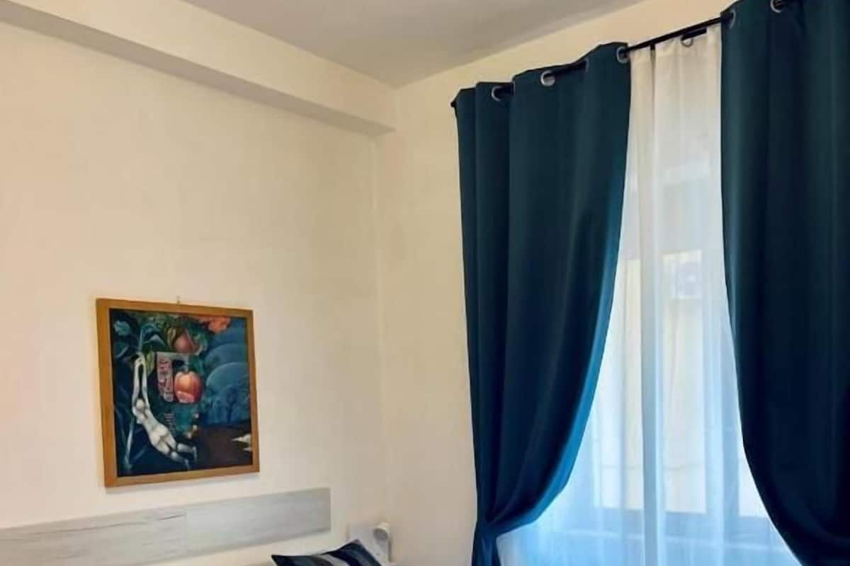 A hotel room with a bed, pillows, a painting on the wall, and a window with curtains.