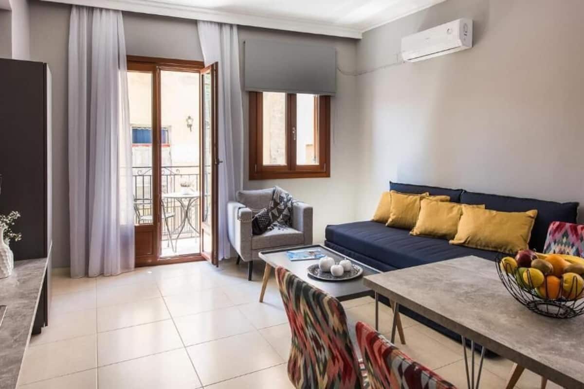 A modern living room with a sofa, a dining table, and a coffee table. There is a balcony with a view of the outside.