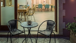 Un espace repas intérieur avec des chaises en osier et une petite table ronde, une table à manger en bois avec des chaises, et une vue sur une pièce dotée d’une grande fenêtre et d’un lustre.