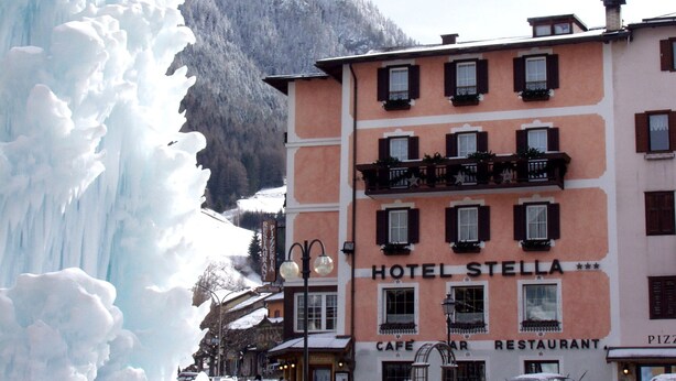 A hotel named 'HOTEL STELLA' with a 'CAFE' and 'RESTAURANT' sign, a wooden fence, and a large ice formation in the background. - Hotel Stella (Moena)