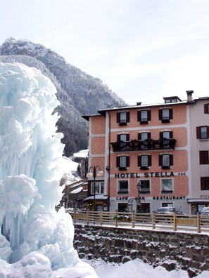 A hotel named 'HOTEL STELLA' with a 'CAFE' and 'RESTAURANT' sign, a wooden fence, and a large ice formation in the background. - Hotel Stella (Moena)