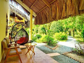 A covered outdoor area with a thatched roof, a hanging chair, a table, and a pathway leading to a lush garden.
