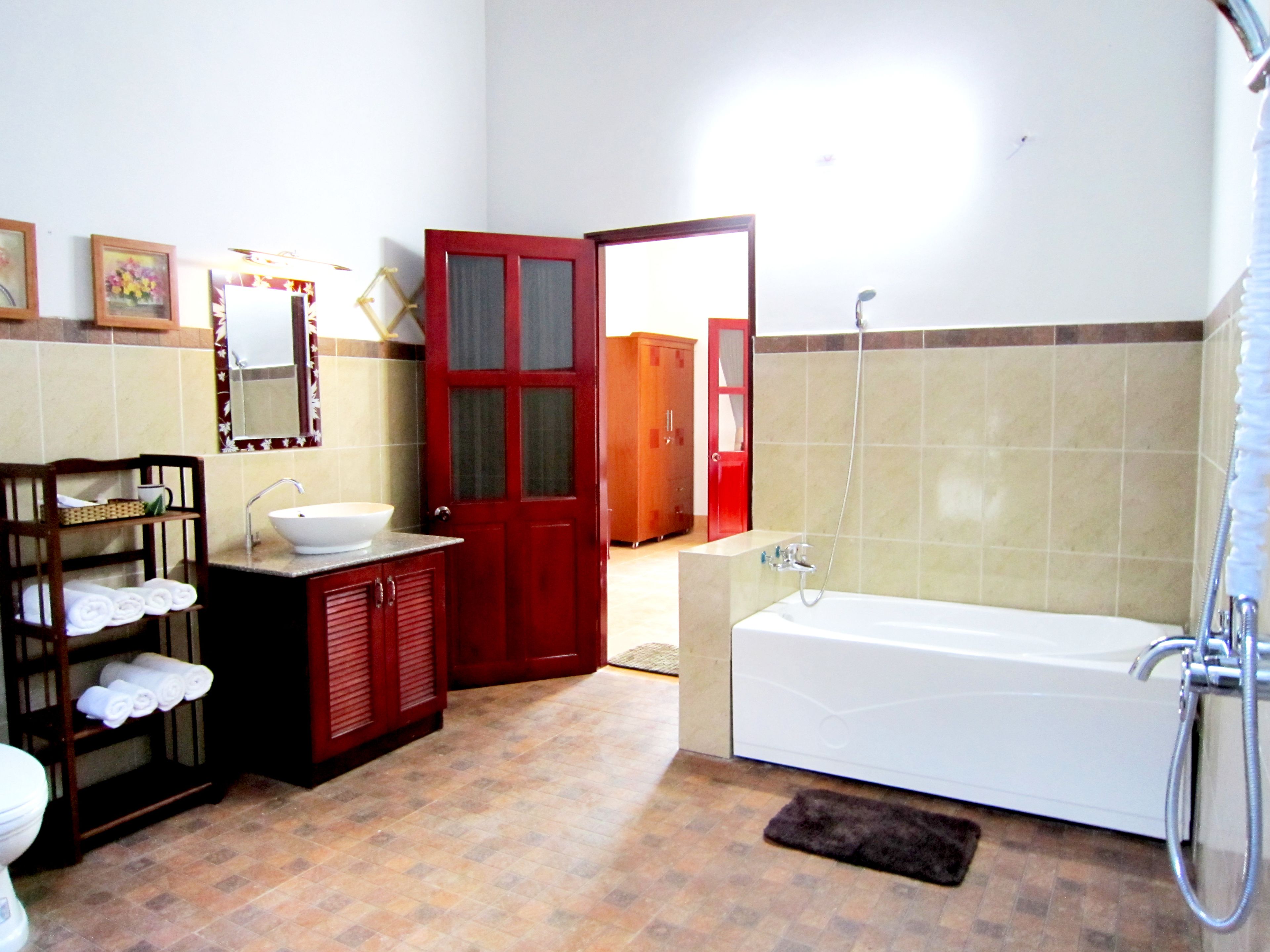 A bathroom with a bathtub, sink, and toilet. There is a wooden cabinet with towels and a mirror above the sink. A red door is visible in the background.