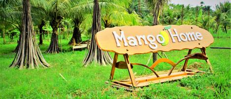 Un letrero de madera con el texto "Mango Home" en un paisaje verde exuberante con palmeras.