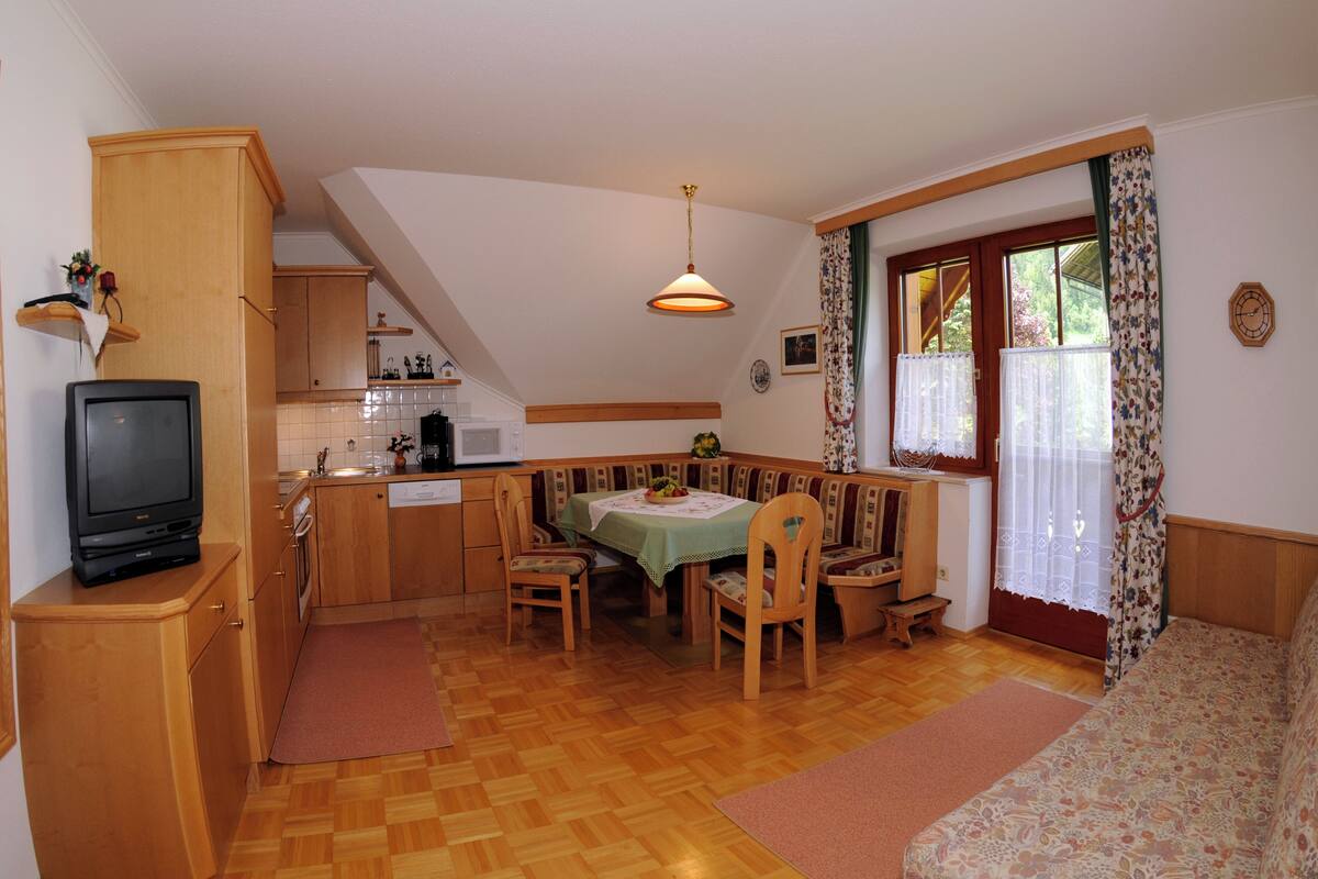 Eine kleine Küche mit Holzschränken, einem Esstisch mit Stühlen, einer eingebauten Bank und einem Fernseher auf einem Schrank.