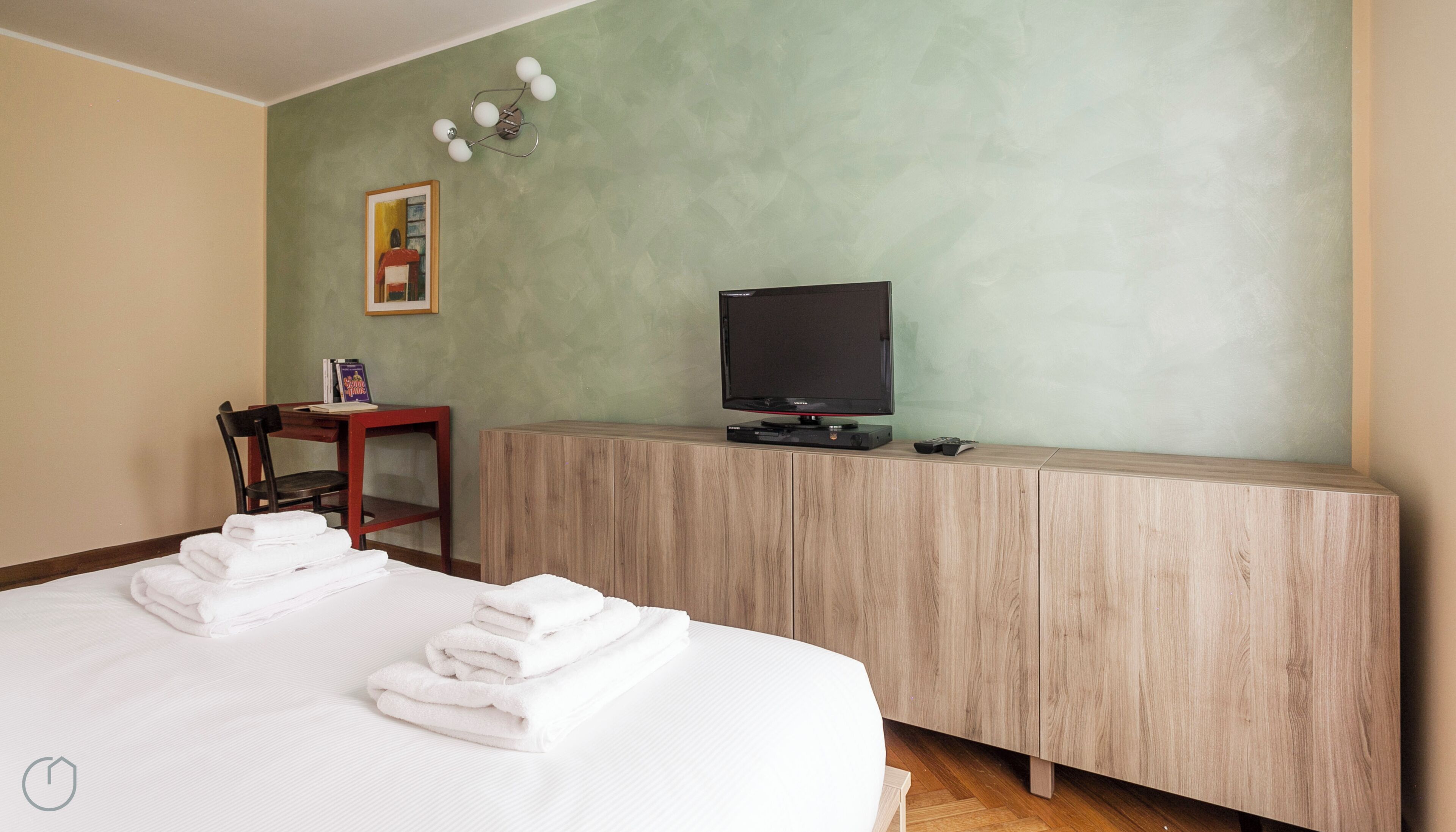Una habitación de hotel con una cama, un televisor sobre una cómoda de madera, una mesita con un libro y un cuadro en la pared.