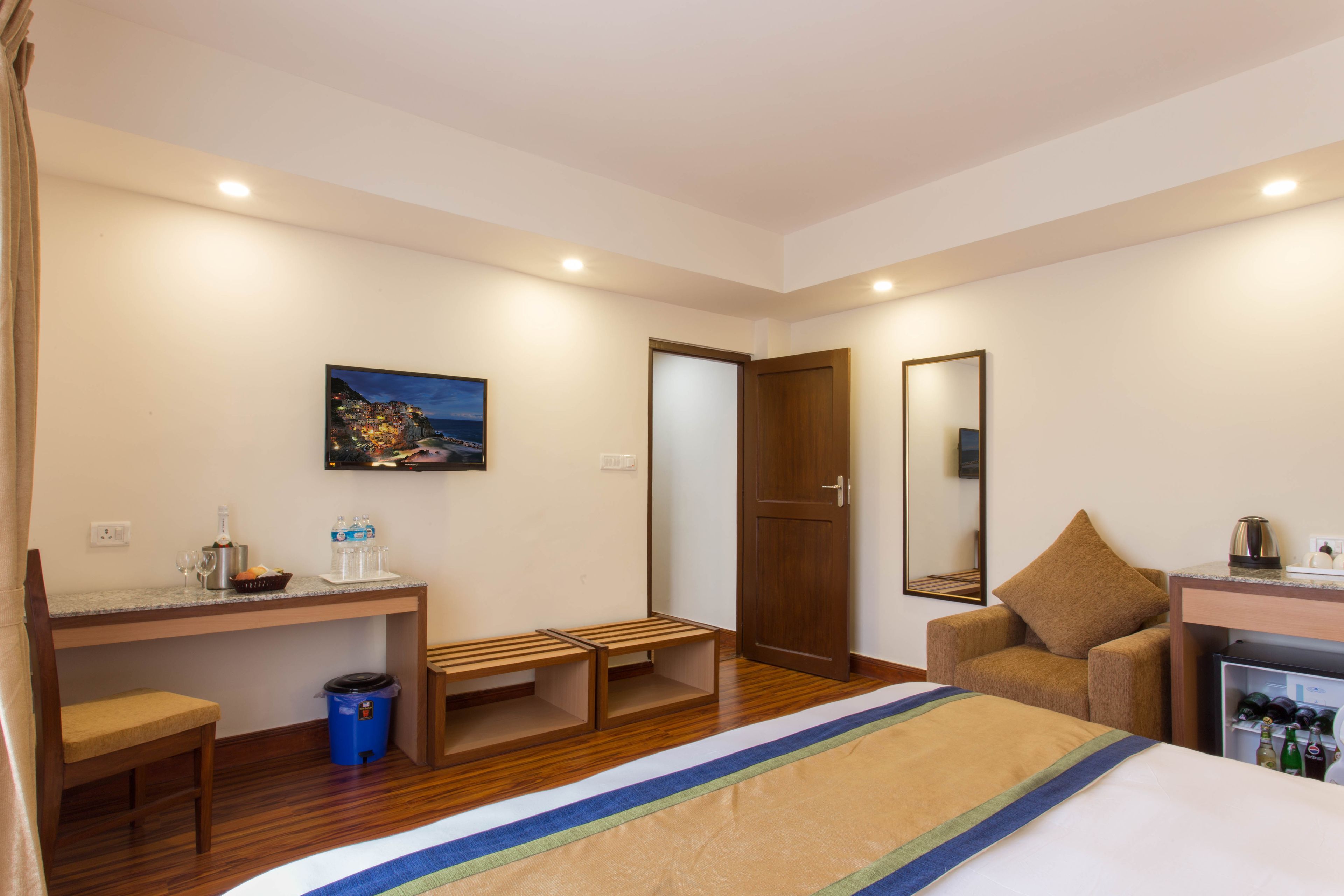 A hotel room with a bed, a desk with a chair, a small table with bottles, a trash bin, a bench, a door, a mirror, and a picture on the wall.