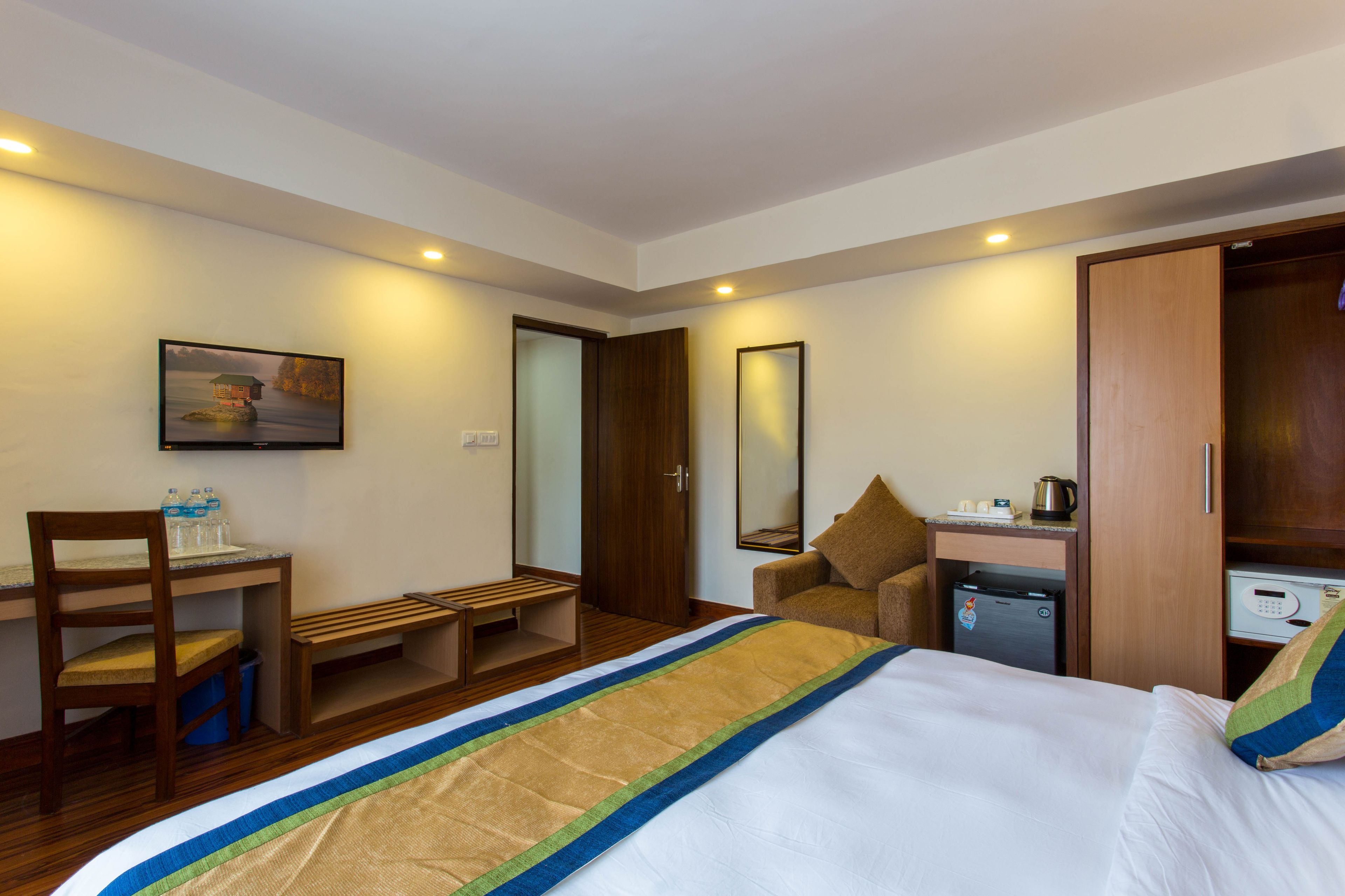 A hotel room with a bed, a desk, a chair, a small table, a mirror, a wardrobe, and a refrigerator.
