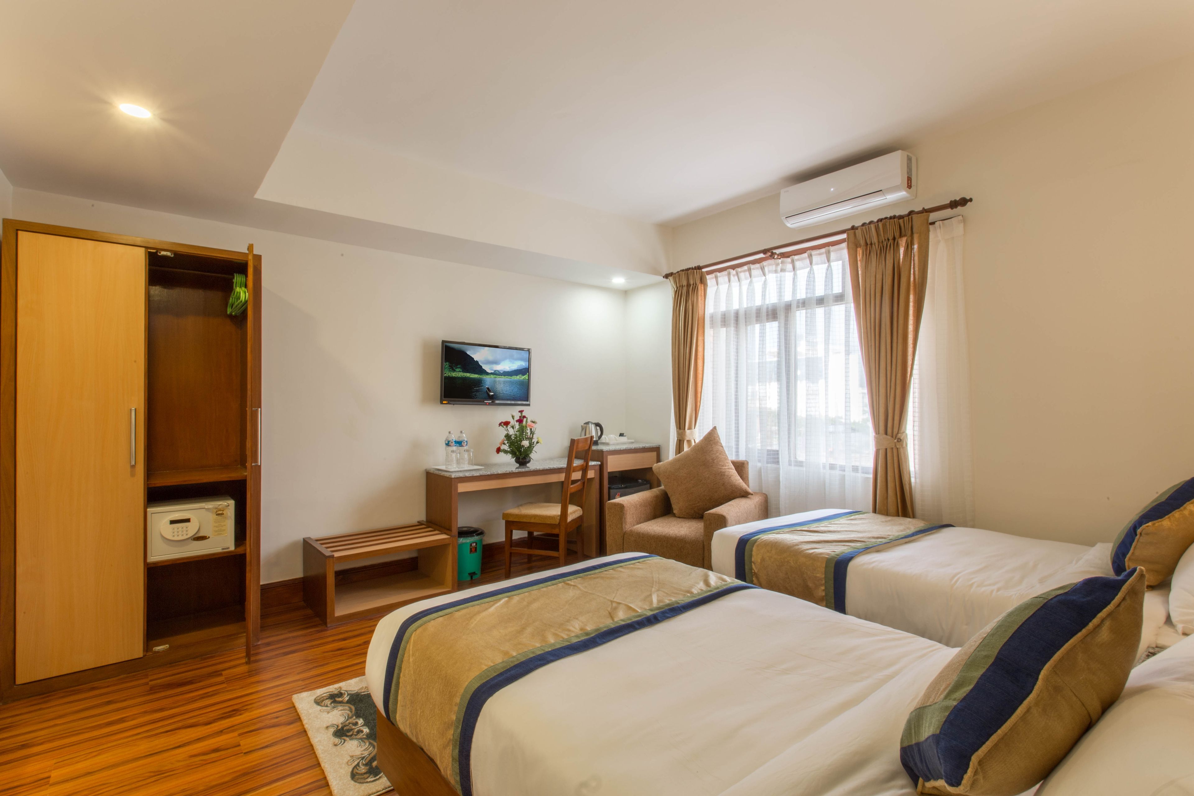 A hotel room with two beds, a desk, a chair, a wardrobe, and an air conditioning unit.