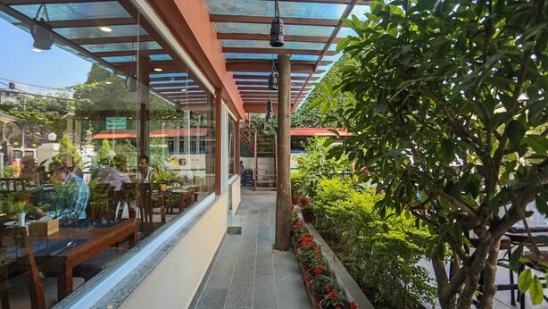 Un café al aire libre con mesas y sillas, una estructura de vidrio y plantas en macetas.