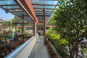 Un café al aire libre con mesas y sillas, una estructura de vidrio y plantas en macetas.