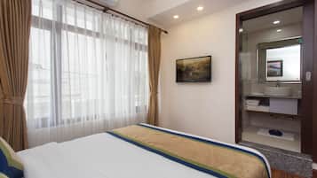 A hotel room with a bed, a large window with sheer curtains, a painting on the wall, and a bathroom with a mirror and sink.