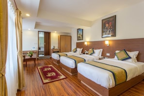 Habitación de hotel con tres camas, piso de madera, alfombra roja, escritorio con silla, espejo y un cuadro en la pared.