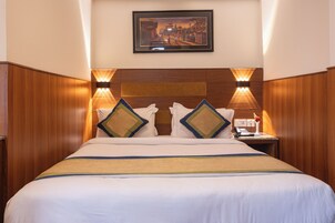 Una habitación de hotel con una cama grande, cabecera de madera, dos lámparas fijadas a la pared, una mesita de noche con un teléfono y un cuadro enmarcado en la pared.