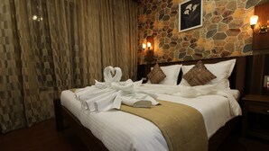 A neatly made bed with white linens and a brown throw blanket, a stone wall with a floral painting, and a wooden nightstand with a lamp. - Hotel Majestique pune (Pune)