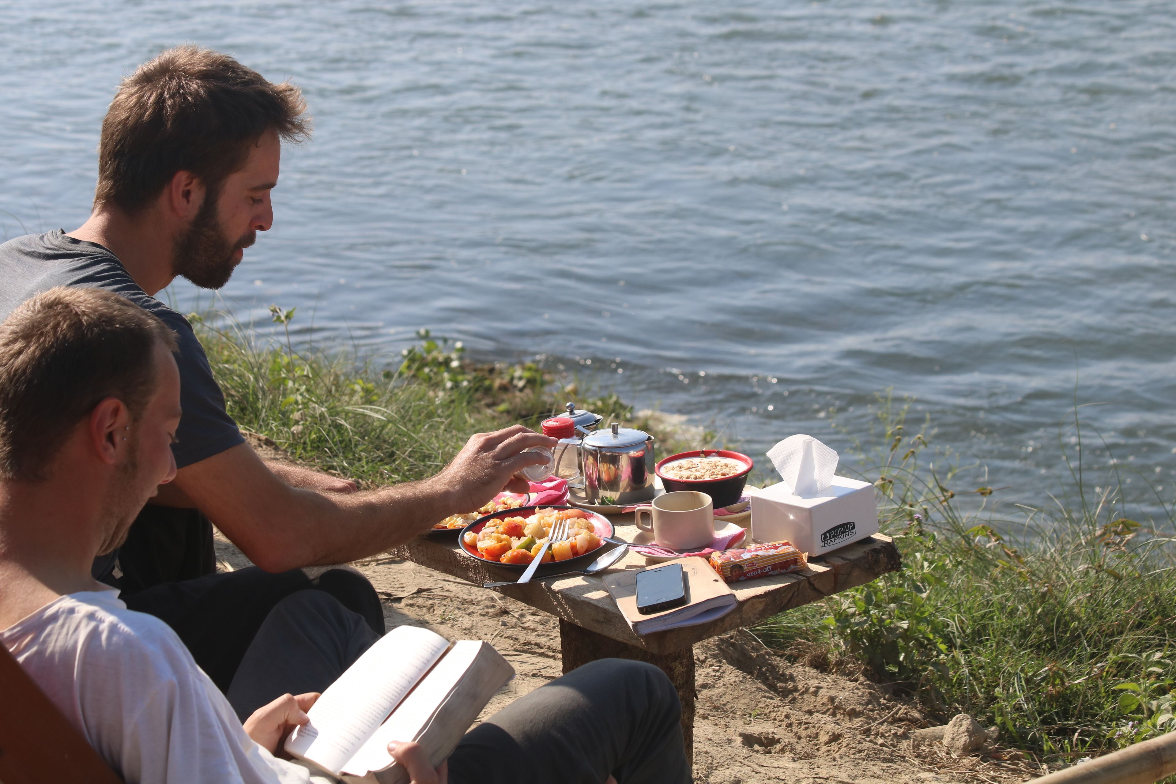 Zwei Personen sitzen an einem Holztisch am See, mit Speisen und Getränken.