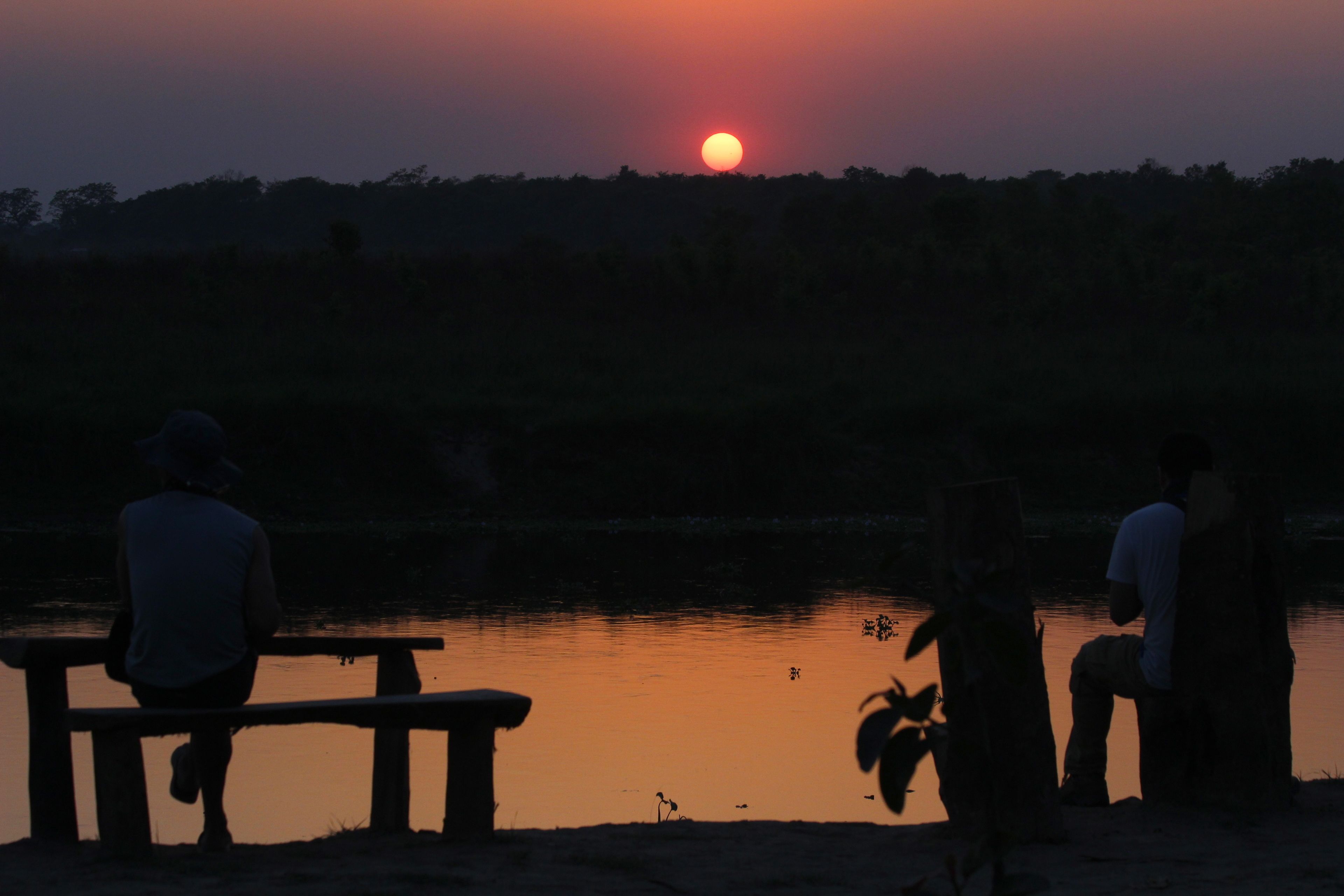 A sunset over a body of water with people sitting on benches.