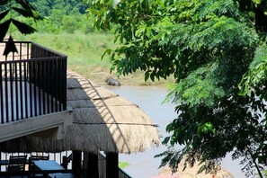 A thatched-roof outdoor area with a wooden deck and tables, overlooking a river. - Jungle Sunset Camp (Sauraha)