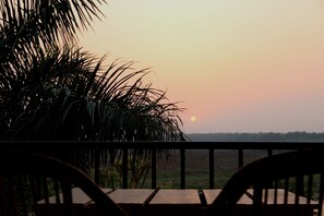 A sunset view from a balcony with a wooden table and chairs, overlooking a grassy landscape. - Jungle Sunset Camp (Sauraha)