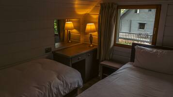 A room with two beds, a small desk with lamps, a window with curtains, and a view of a house outside.