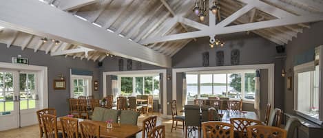 Une salle à manger spacieuse, dotée de tables et de chaises en bois, de grandes fenêtres offrant une vue sur l’extérieur et d’un plafond haut avec des poutres apparentes.