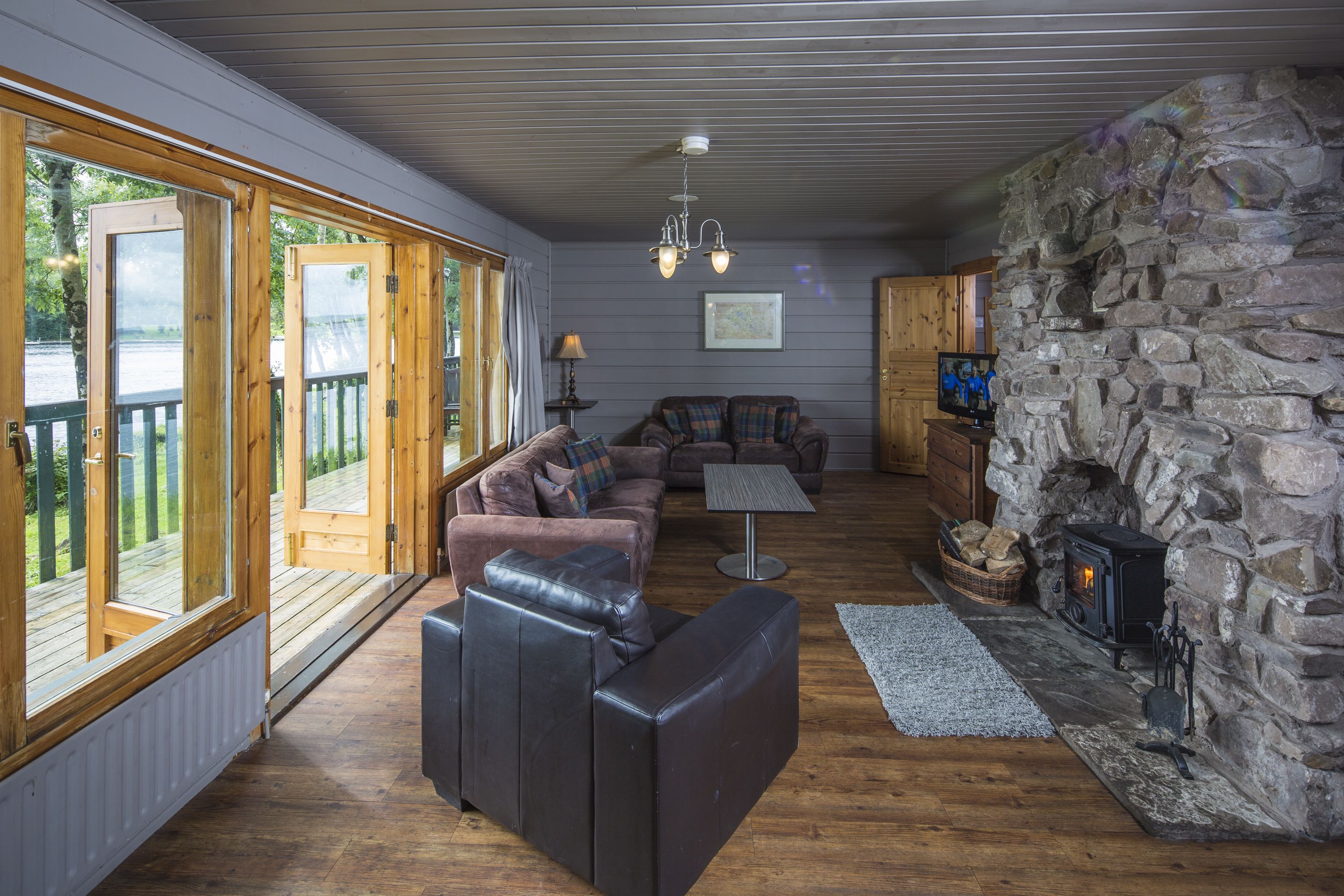 A cozy living room with a stone fireplace, leather sofas, a television, and large windows overlooking a deck.