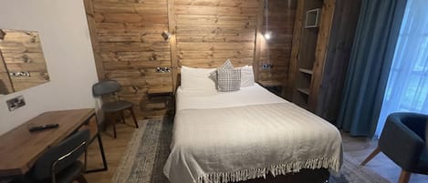 A hotel room with a wooden wall, a bed with a white bedspread, a desk with a chair, and a wardrobe.
