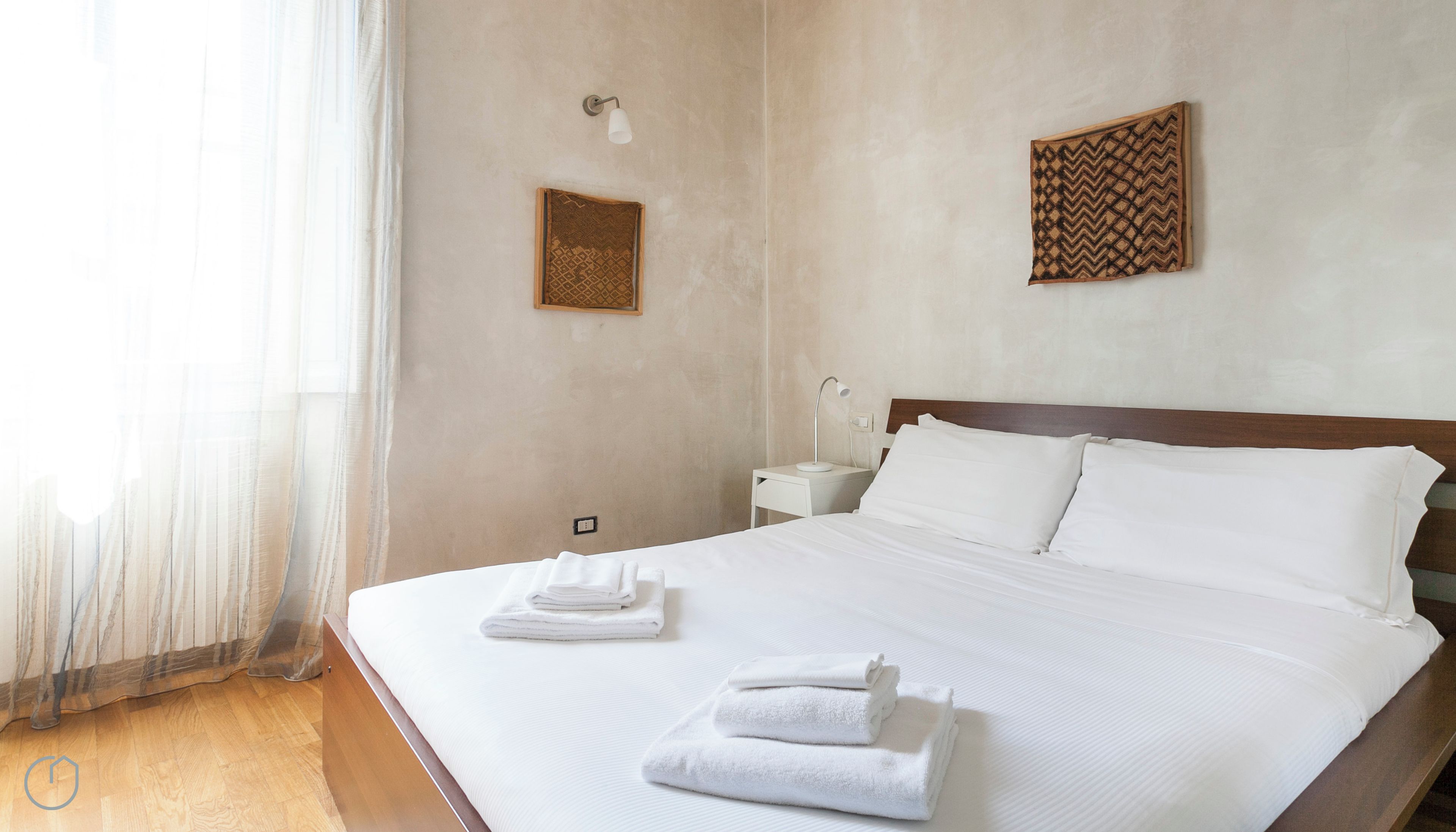 Habitación de hotel con una cama grande, ropa de cama blanca, una mesita de noche con lámpara y una lámpara de pared.