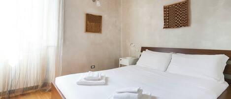 Habitación de hotel con una cama grande, ropa de cama blanca, una mesita de noche con lámpara y una lámpara de pared.