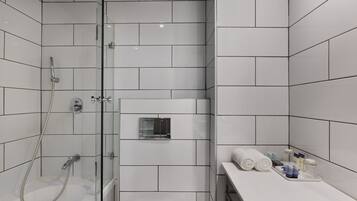 Un baño moderno con azulejos blancos, cabina de ducha de vidrio, tocador con artículos de aseo e igual un espejo pequeño.